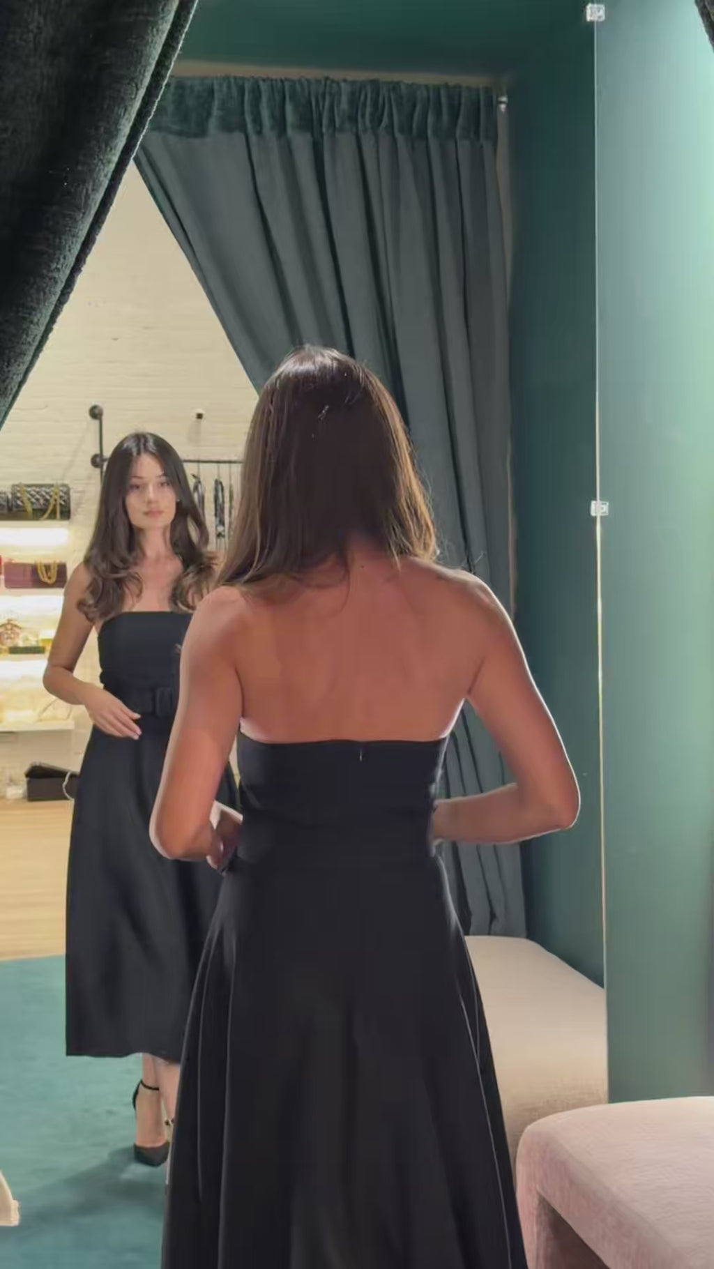 A woman trying on a premium black strapless cocktail dress in a fitting room. She adjusts the belt, showcasing the flawless architectural fit and structured fabric ideal for black tie events.
