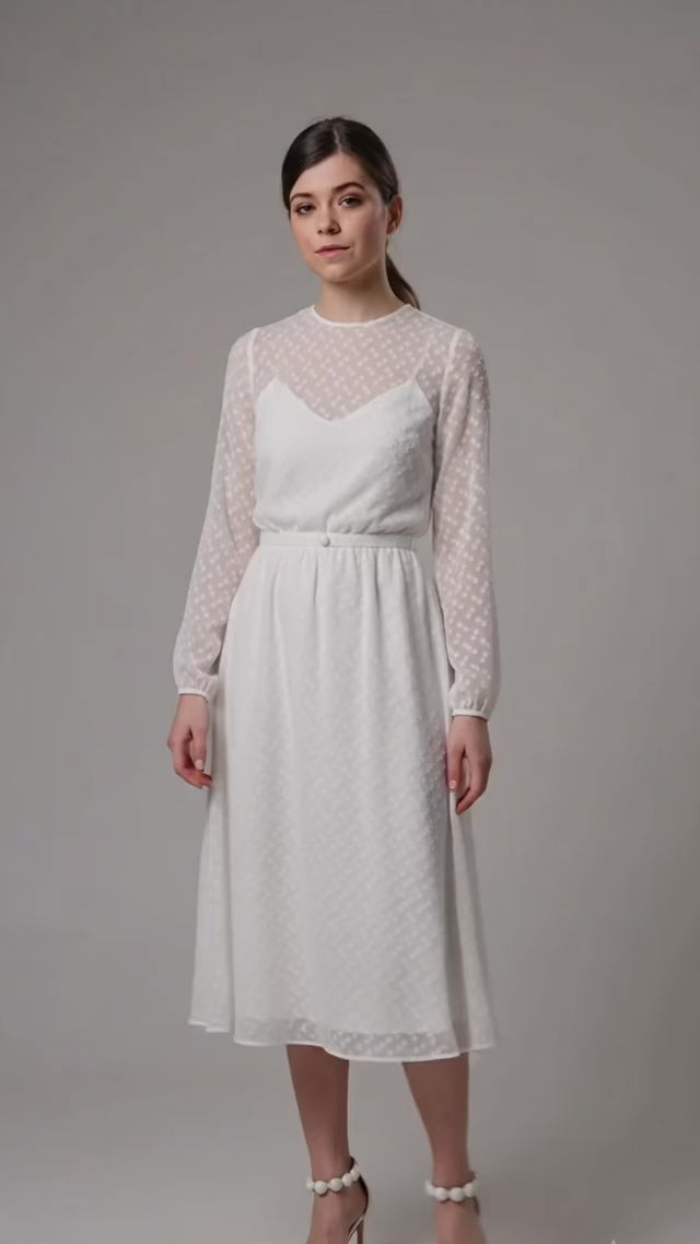 Video of a model twirling in an elegant white midi dress by Anastasiia Ivanova. The dress features a delicate sheer dotted overlay, long sleeves, and a flowy skirt, showcasing a flawless look perfect for a civil wedding or rehearsal dinner